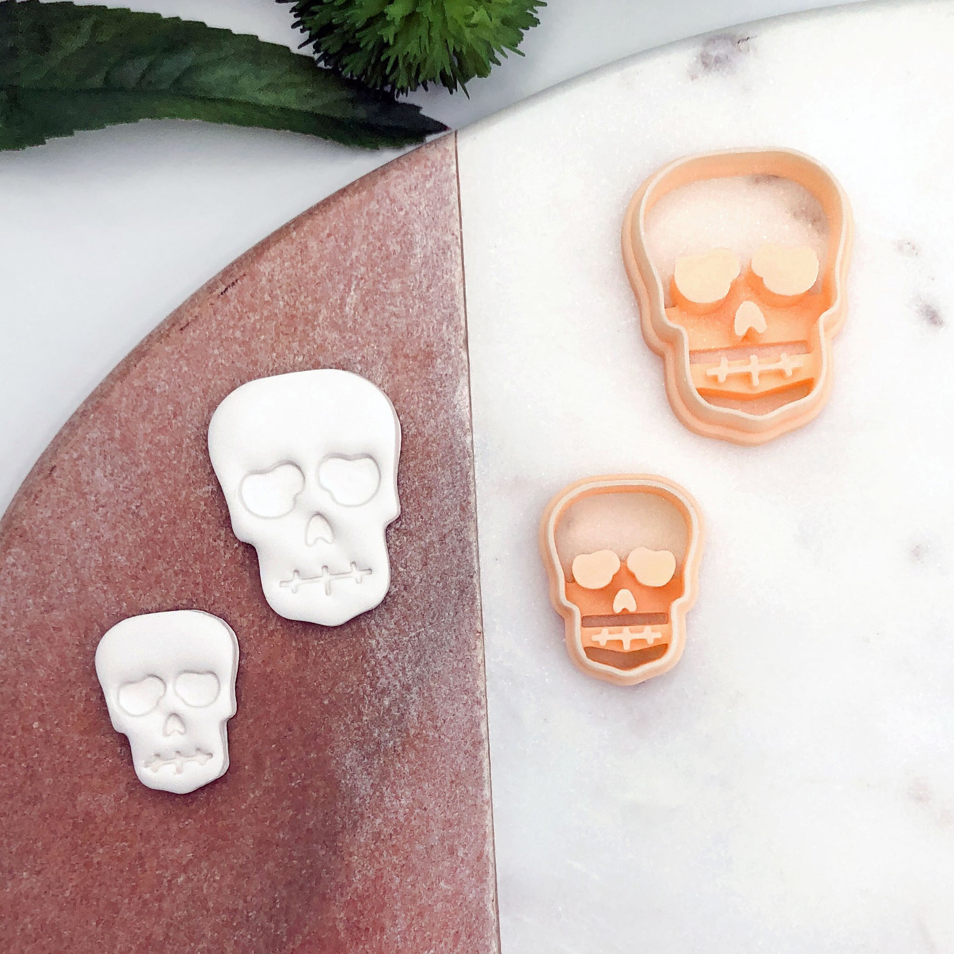 Two orange plastic skull-shaped cookie cutters displayed on a white marble surface next to a rose-gold tray holding two white skull-shaped cutouts, with a green leaf in the top-left corner.