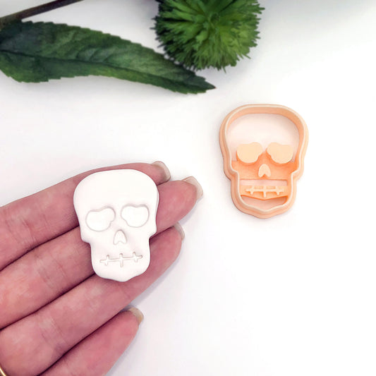 A white skull-shaped item held in a person's hand next to a peach-colored matching  cookie cutter on a white background, with green decorative leaves in the upper corner.