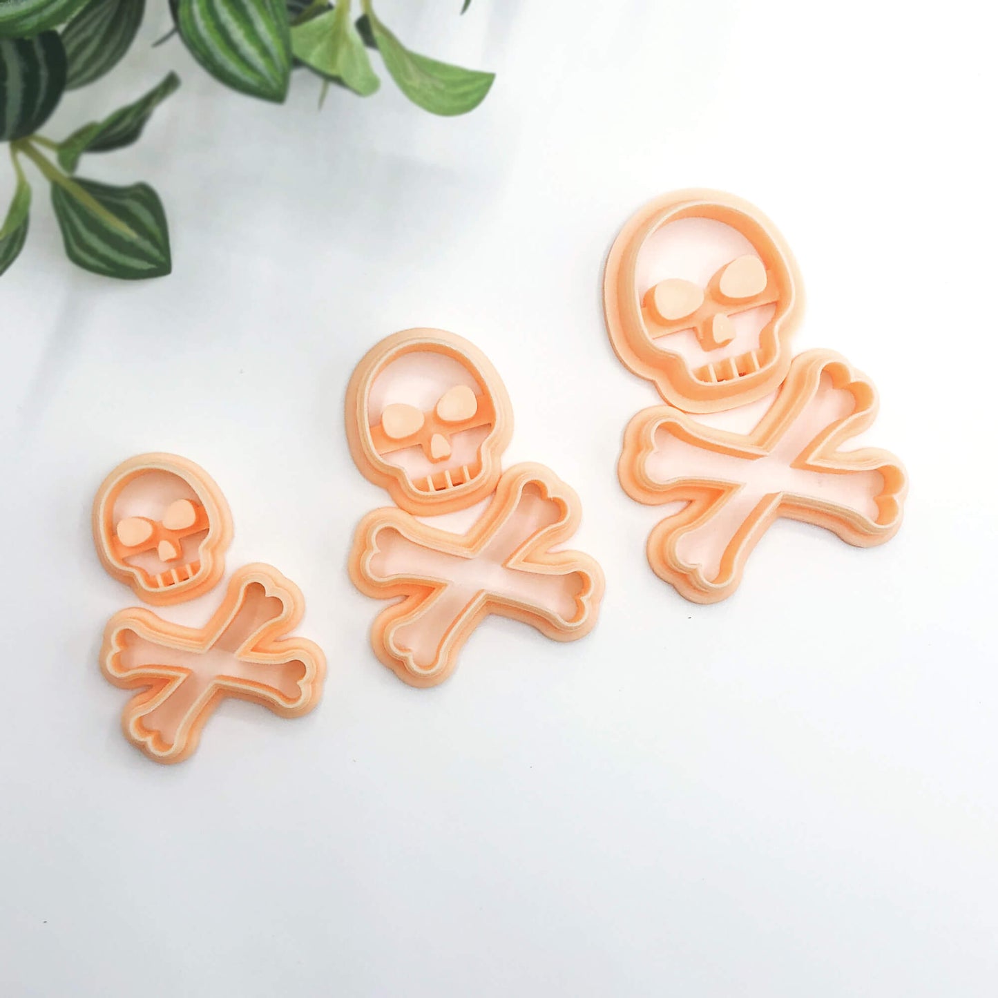 Three peach-colored plastic skull and crossbones cookie cutter in different sizes, arranged diagonally on a white surface with green leaves in the top-left corner.