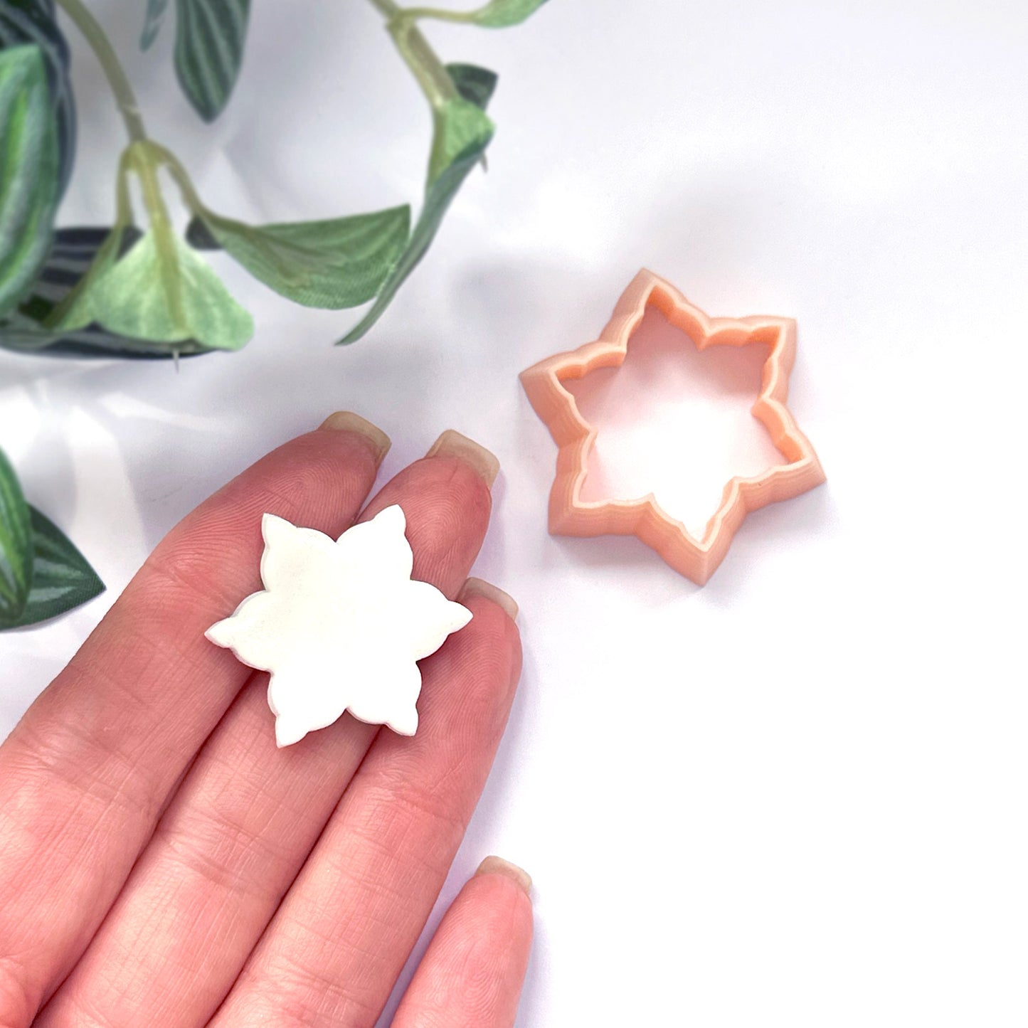 A hand holds a small white snowflake-shaped polymer clay cut out, with a peach-colored snow star-shaped cutter placed nearby on a white surface. Green leaves are visible in the upper left corner.