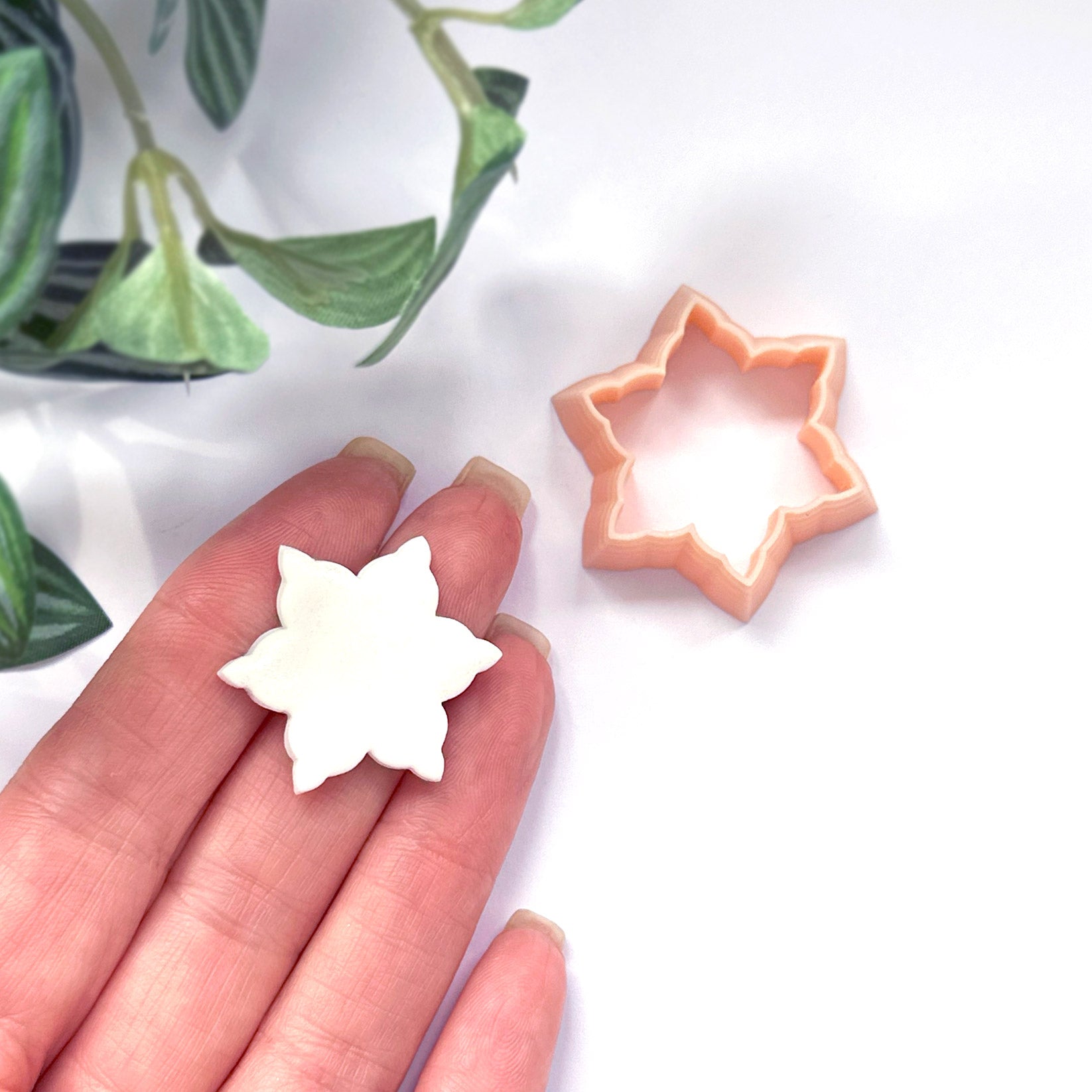 A hand holds a small white snowflake-shaped polymer clay cut out, with a peach-colored snow star-shaped cutter placed nearby on a white surface. Green leaves are visible in the upper left corner.