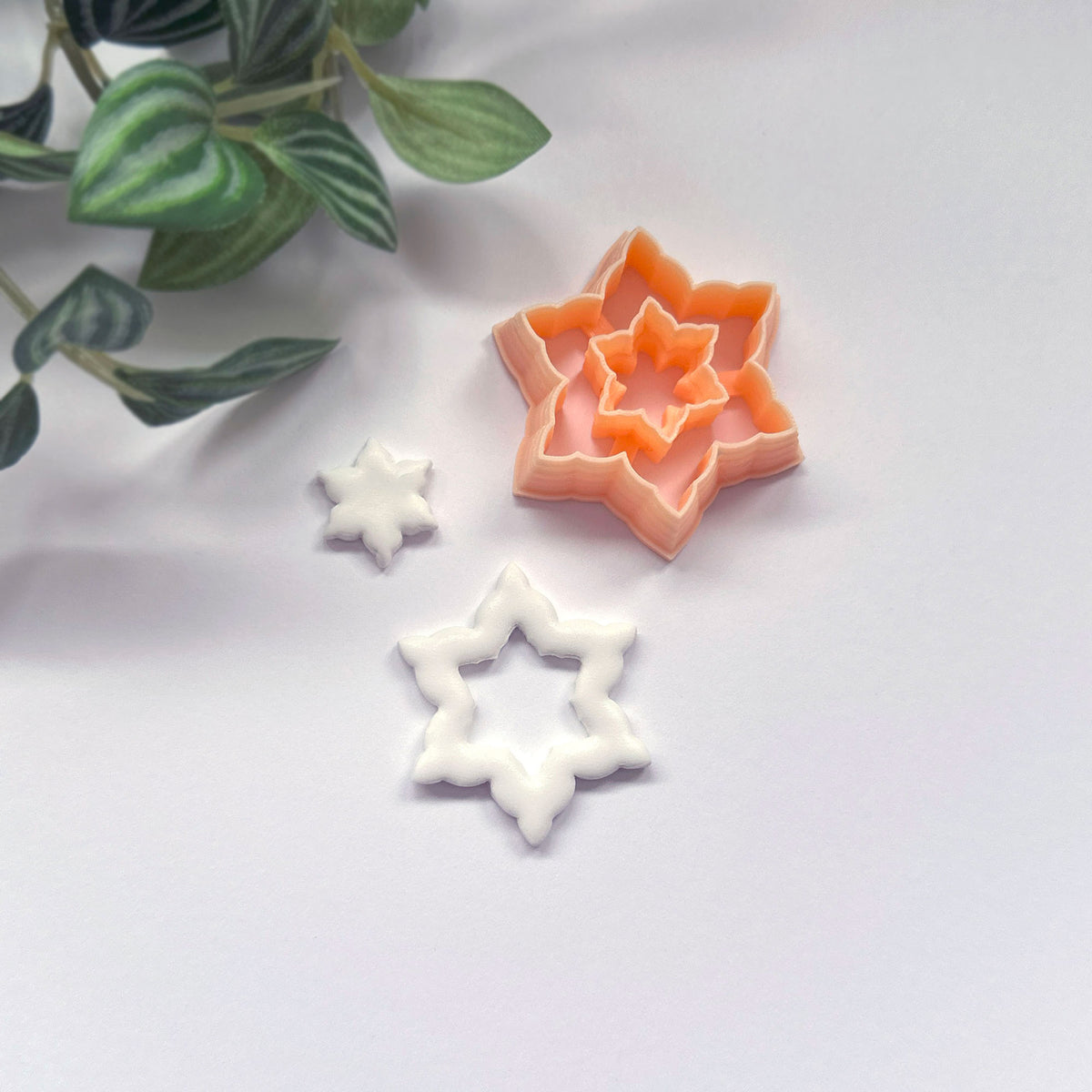 A snowflake star-shaped cookie clay cutter in peach isdisplayed on a light gray surface with a decorative green plant in the corner, a white cut out sits beside it.