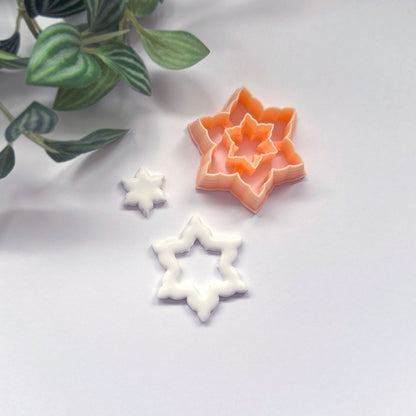 A snowflake star-shaped cookie clay cutter in peach isdisplayed on a light gray surface with a decorative green plant in the corner, a white cut out sits beside it.