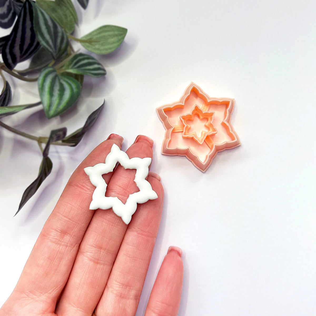 A hand holds a small white star-shaped snowflake clay cutout while a peach-colored star snowflake-shaped cookie clay cutter sits on a white surface, with green plant leaves in the upper left corner.