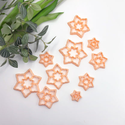 A set of peach-colored plastic cookie clay cutters in various snowflake starburst shapes, arranged on a white surface with green leaves in the top-left corner.