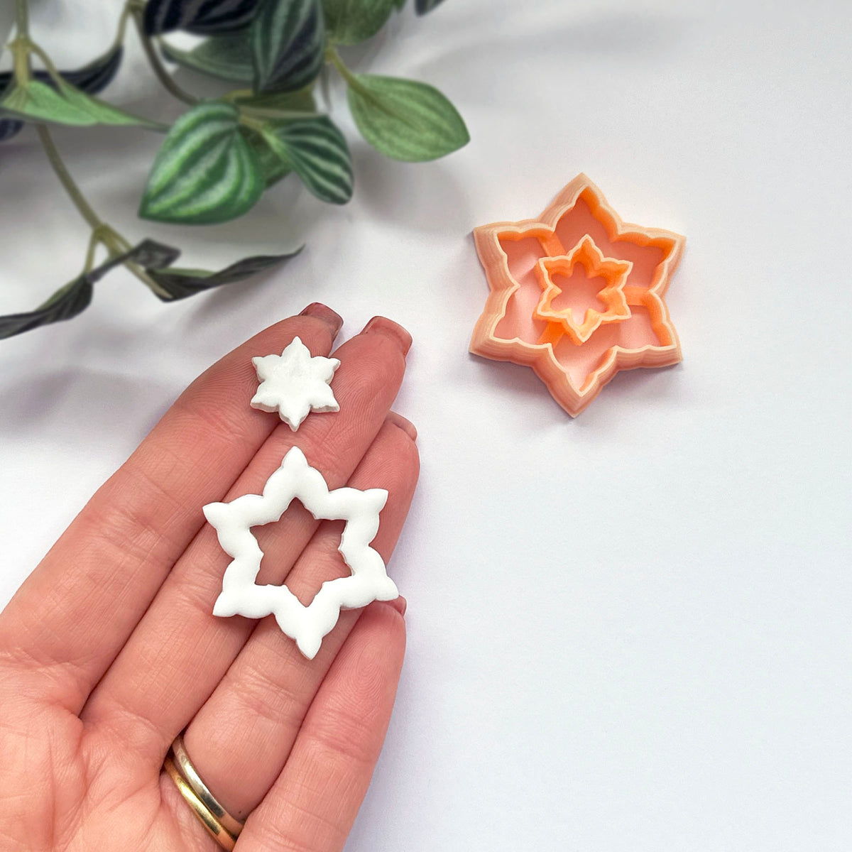 A hand holds two small white snowflake-shaped earring cutouts, with a peach-colored star-shaped cookie cutter placed on a white surface in the background. Green leaves are visible in the upper left corner.