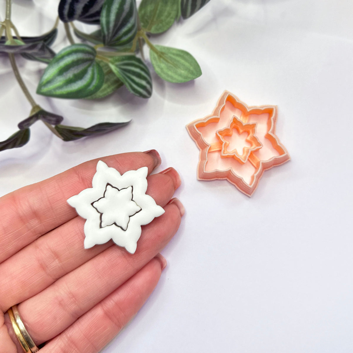 A hand holds a white star snowflake-shaped cut out, with a matching peach star-shaped cutter placed beside it on a white surface. Green leaves are visible in the upper left corner.