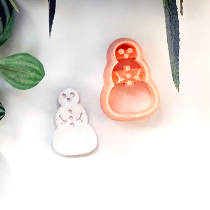 A small snowman-shaped white polymer clay cut out placed beside a peach-colored snowman-shaped cutter on a white surface with decorative green leaves at the edges.