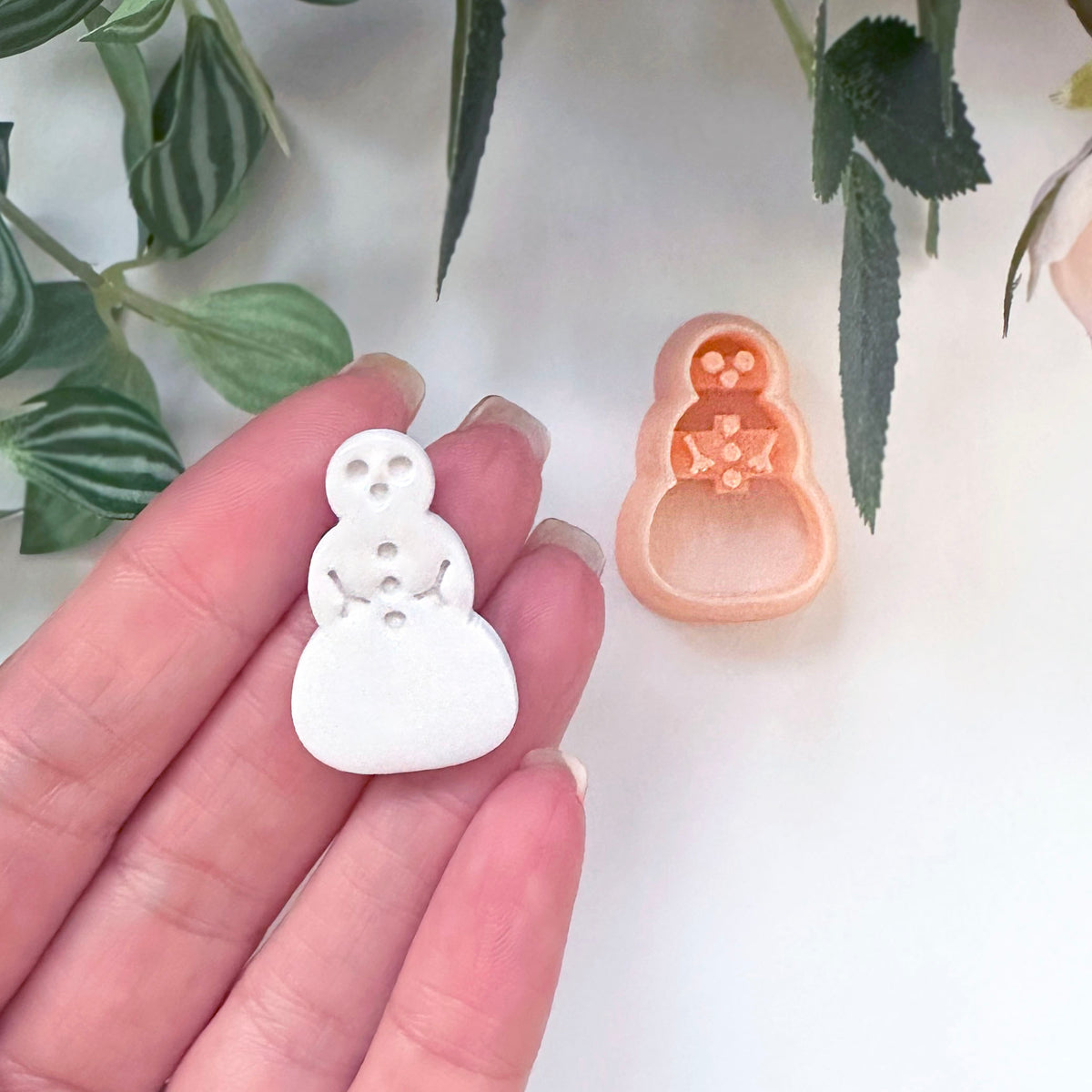A hand holds a small white snowman-shaped polymer clay cut out, with a peach-colored matching clay cutter placed beside it on a light surface, with green leaves in the background.