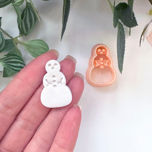 A hand holds a small white snowman-shaped polymer clay cut out, with a peach-colored matching clay cutter placed beside it on a light surface, with green leaves in the background.