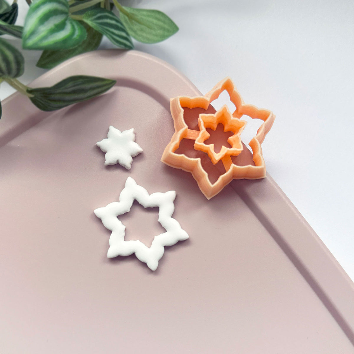 A small white star-shaped snowflake clay cutout for earring design, while a peach-colored star snowflake-shaped cookie clay cutter sits on a white surface, with green plant leaves in the upper left corner.