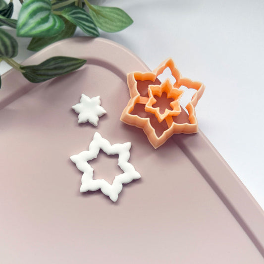 A small white star-shaped snowflake clay cutout for earring design, while a peach-colored star snowflake-shaped cookie clay cutter sits on a white surface, with green plant leaves in the upper left corner.
