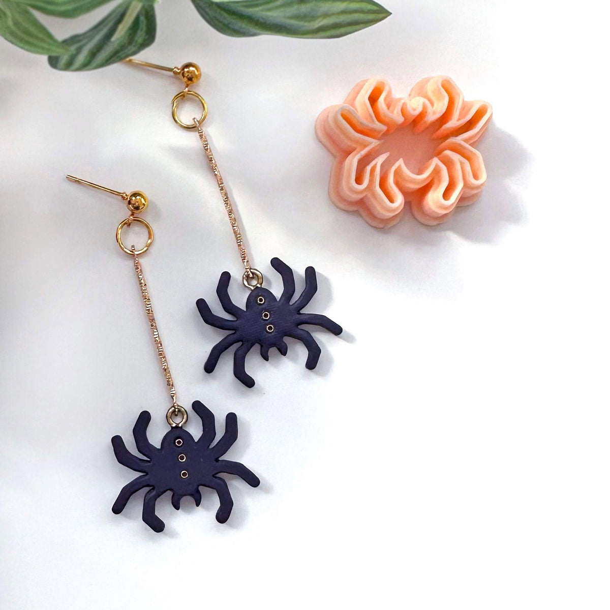 A pair of dangling spider earrings with gold-tone posts and chains, featuring dark navy spider charms, styled on a white background with a peach spider-shaped clay cutter and green leaves in the corner.