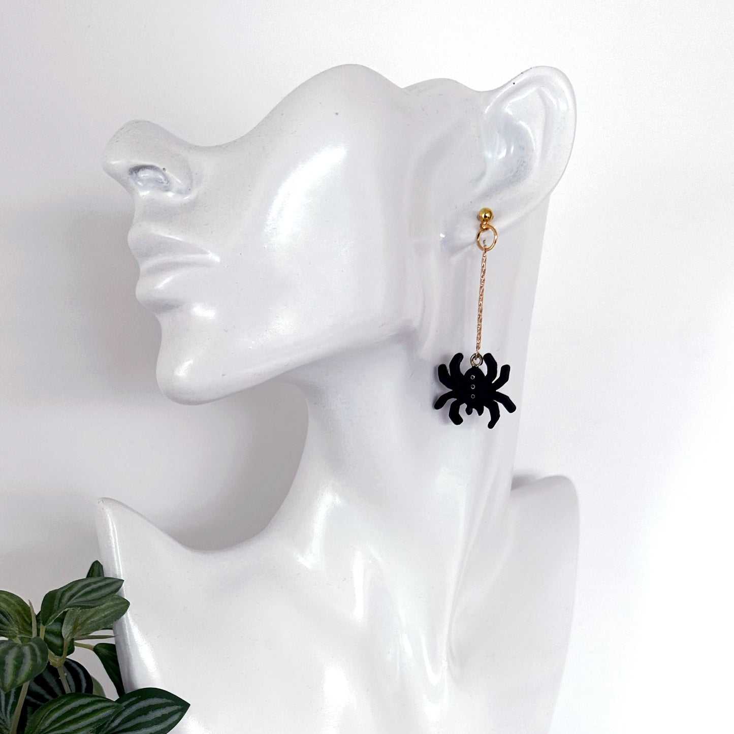 A single dangling earring displayed on a glossy white mannequin head. The earring has a small gold-toned hoop and chain with a black spider charm hanging at the end.
