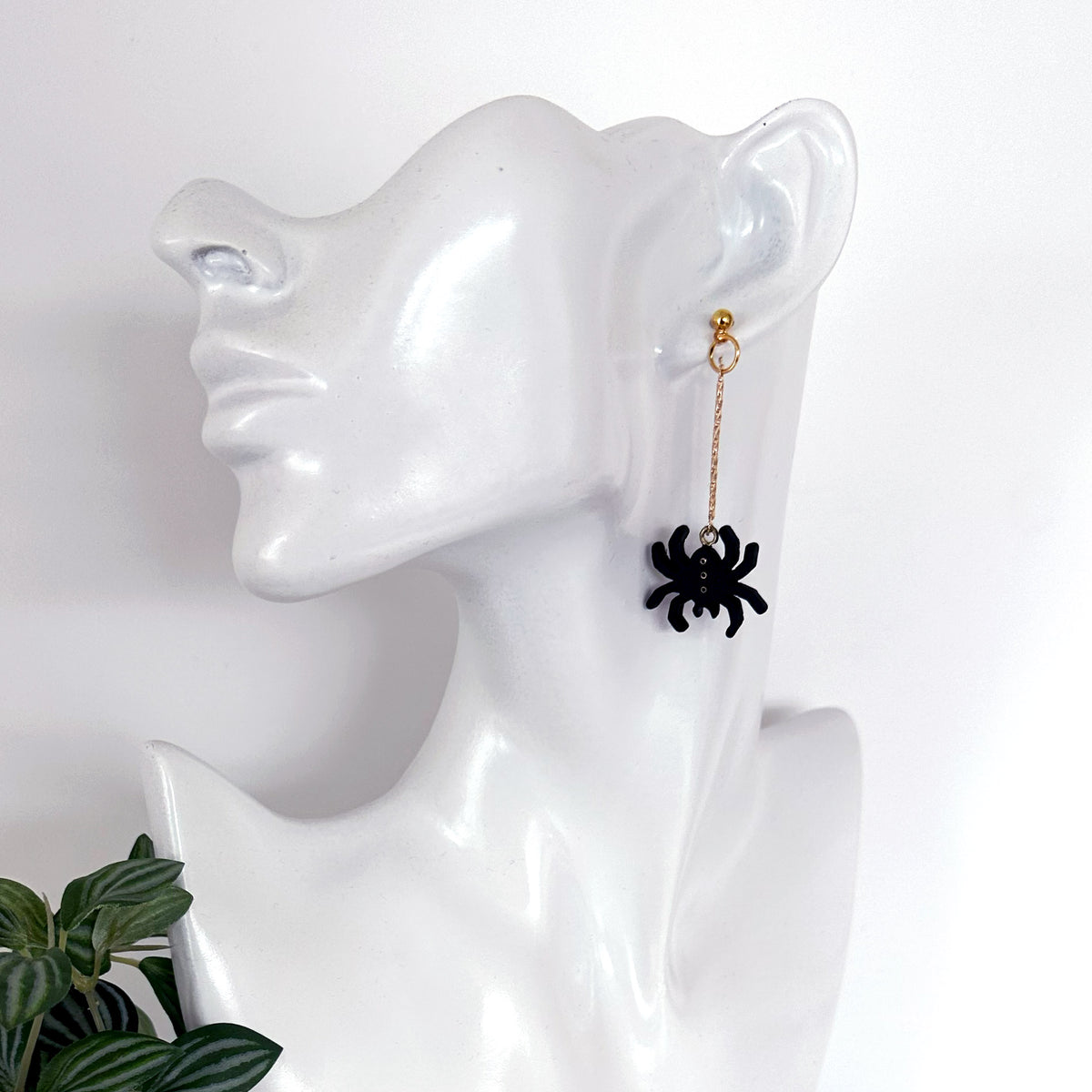 A single dangling earring displayed on a glossy white mannequin head. The earring has a small gold-toned hoop and chain with a black spider charm hanging at the end.