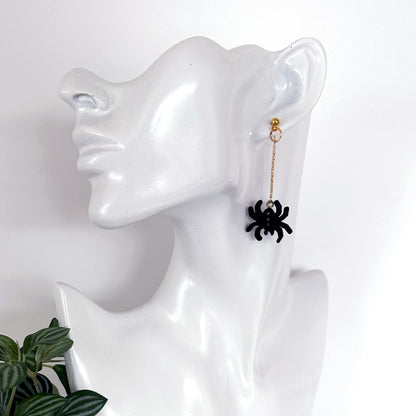 A single dangling earring displayed on a glossy white mannequin head. The earring has a small gold-toned hoop and chain with a black spider charm hanging at the end.