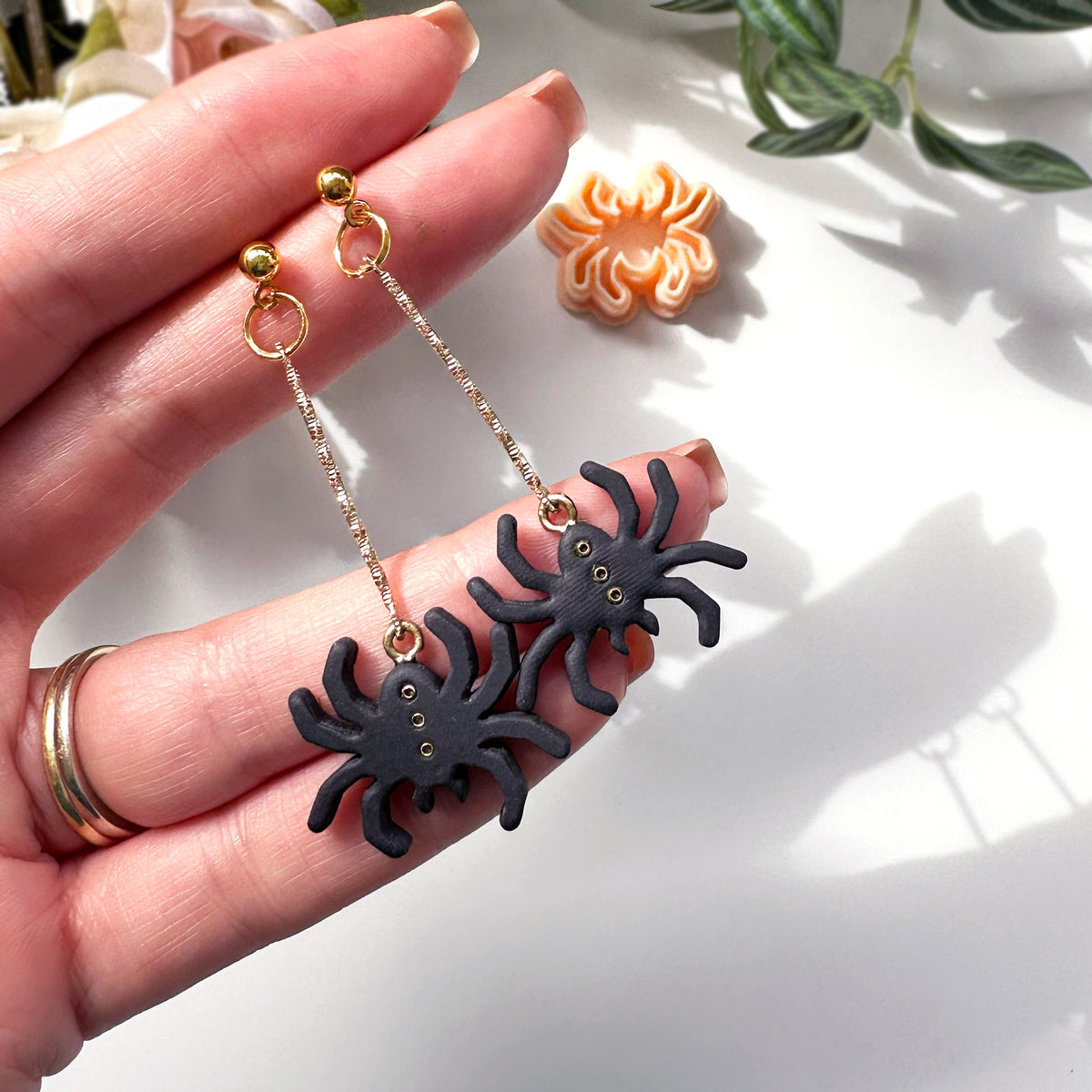 A pair of dangling spider earrings held in a person's hand. The earrings feature black spider charms hanging from delicate gold-tone chains with gold post backings.