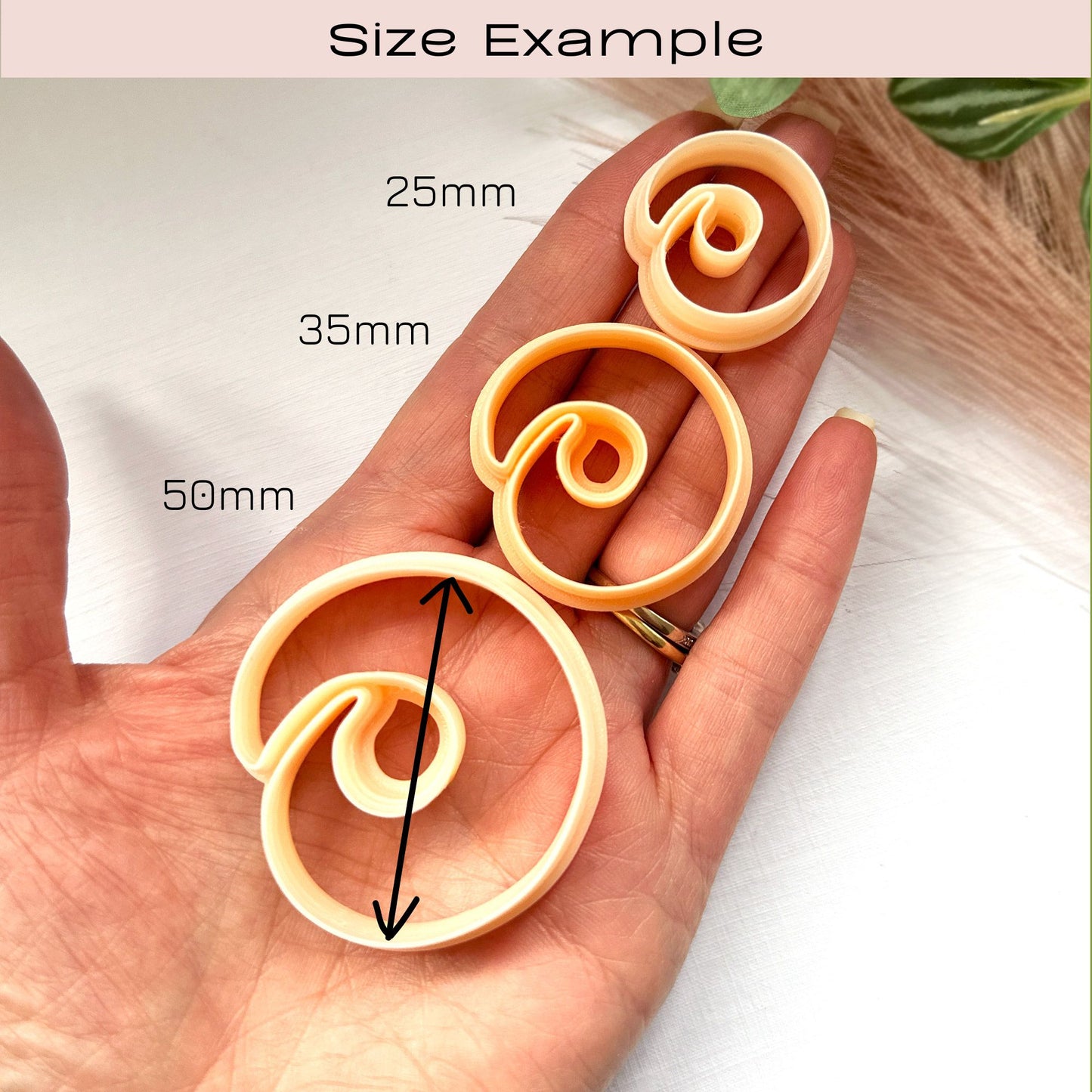 Peach-colored spiral stone clay cutters, for polymer clay earring making -size guide - on a white surface with a plant in the background