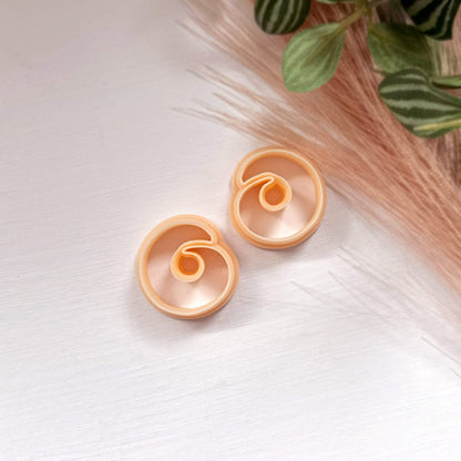 Pair of peach-colored spiral stone clay cutters, mirror pair for polymer clay earring making on a white surface with a plant in the background