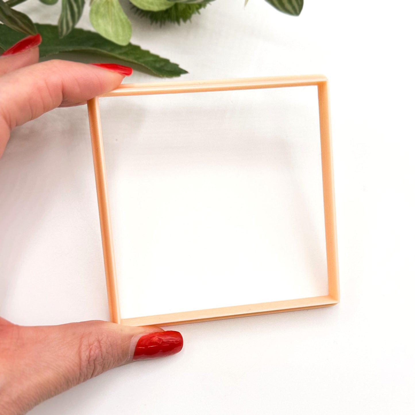 A hand with red-painted nails holds a large square clay cutter against a white background with green leaves in the corner.