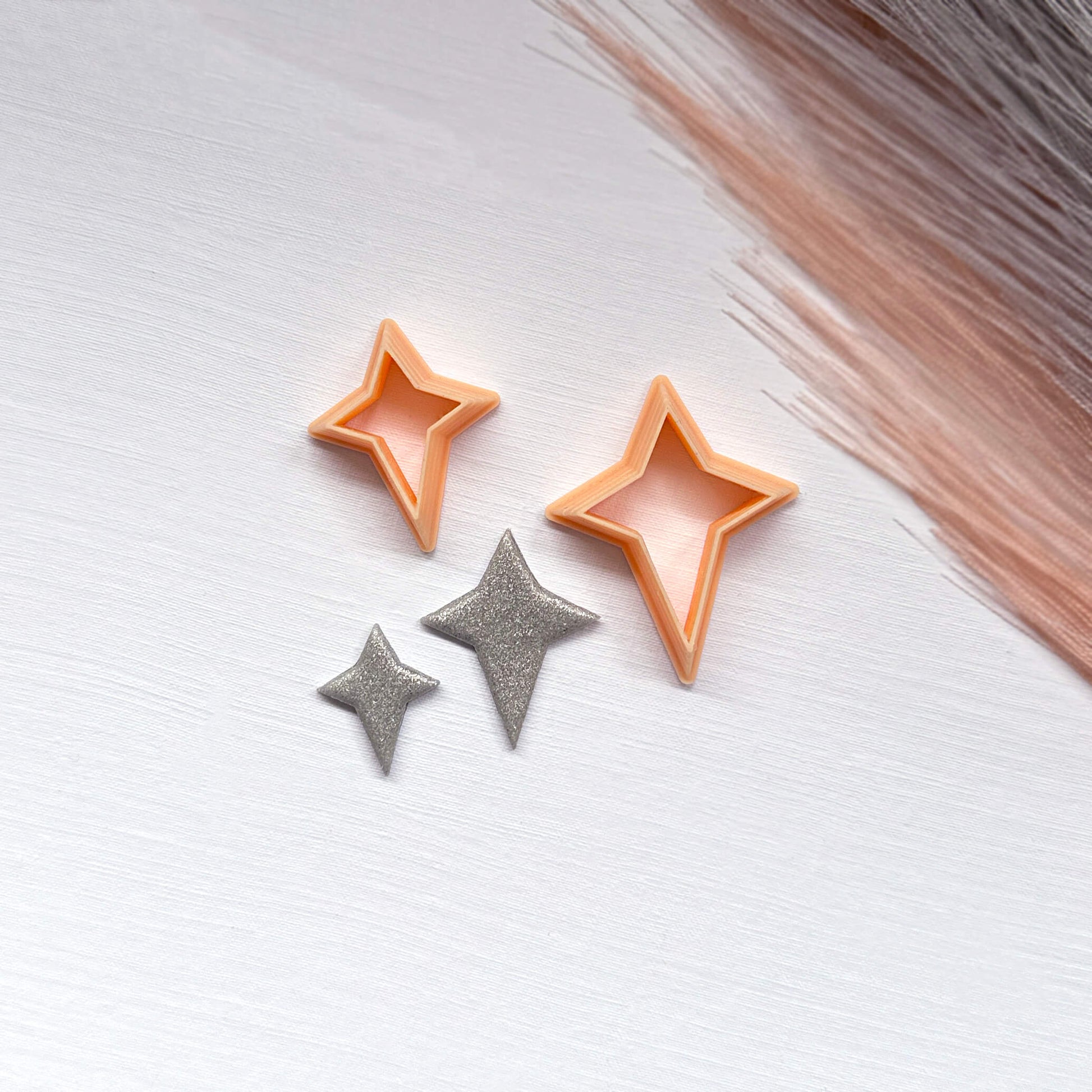 Two small glittery star-shaped clay cutouts next to two star-shaped cookie clay cutters on a light surface.