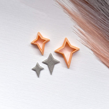 Two small glittery star-shaped clay cutouts next to two star-shaped cookie clay cutters on a light surface.