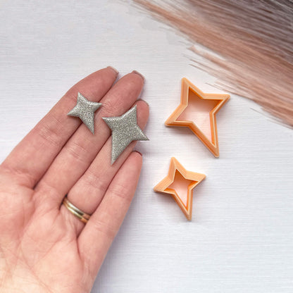 A hand holds two small glittery star-shaped clay cut outs next to two star-shaped cookie clay cutters on a light surface.