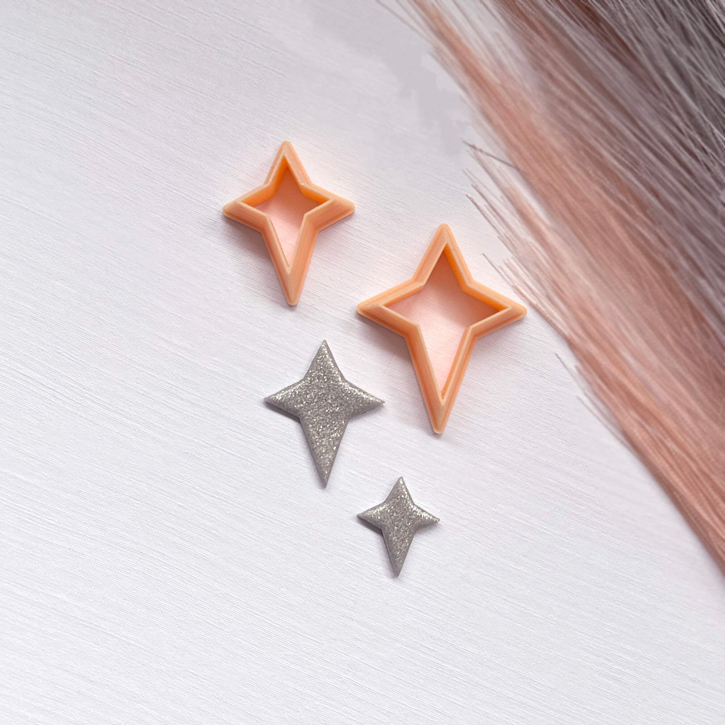 Two small glittery star-shaped clay cutouts next to two star-shaped cookie clay cutters on a light surface.