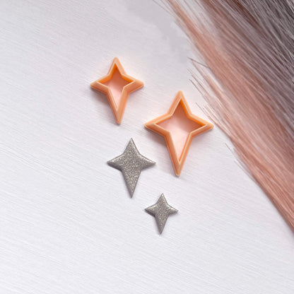 Two small glittery star-shaped clay cutouts next to two star-shaped cookie clay cutters on a light surface.
