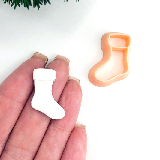 A hand holds a small white Christmas stocking-shaped polymer clay cut out, with a peach-colored matching clay cutter placed on a white surface nearby.
