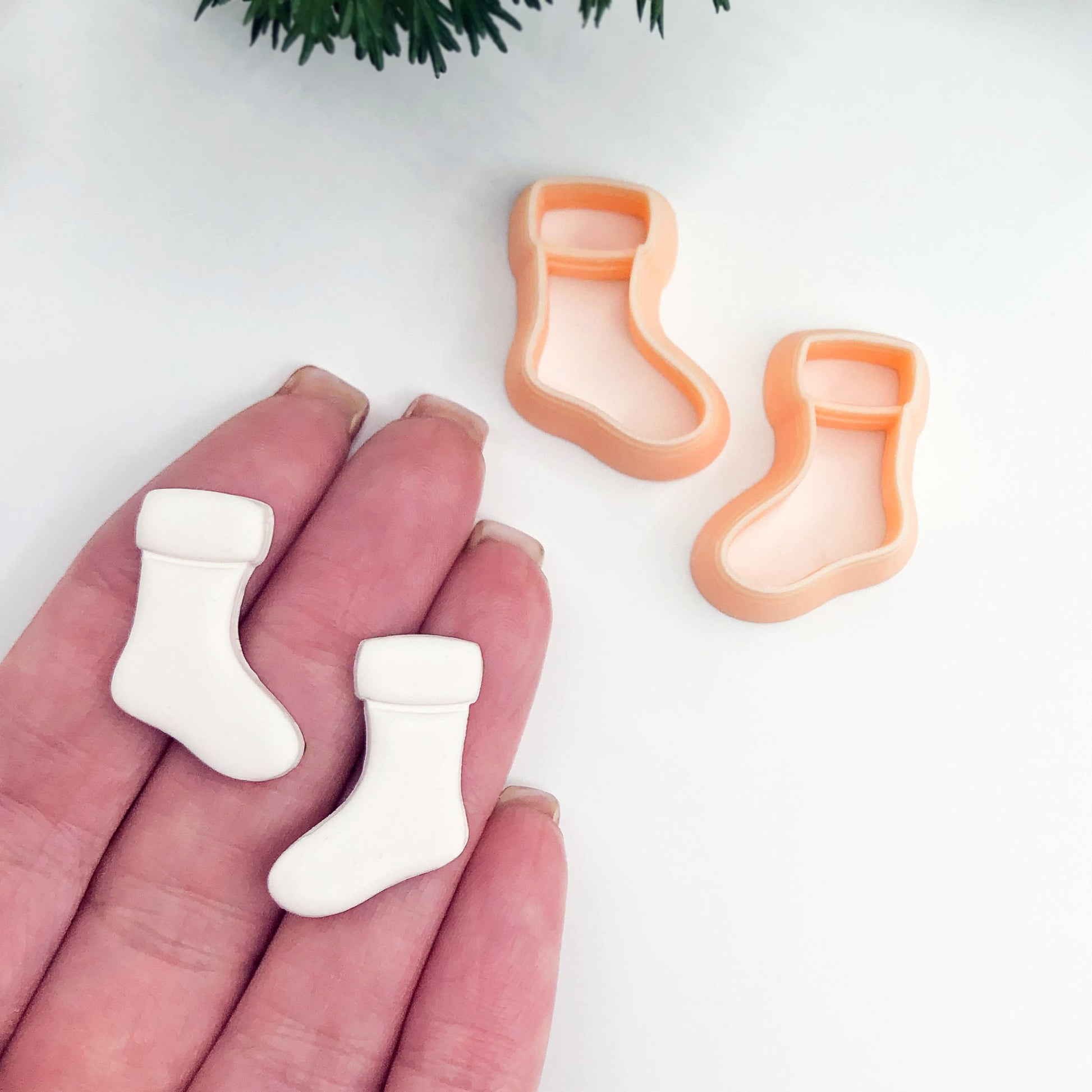 Two small white sock-shaped polymer clay cut outs resting on a person's fingers, with two peach-colored sock-shaped cookie cutters on a white surface behind them.