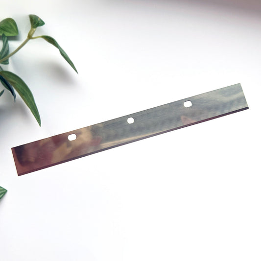 A long metal blade strip, photographed diagonally on a white background with a small green plant visible in the upper left corner.