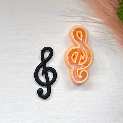 Two treble clef-shaped items on a white textured surface: one matte black clay cutout and one peach-colored plastic clay cutter, with dried grass and a green leaf in the top right corner.