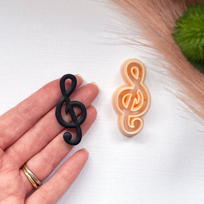 A hand holds a black treble clef-shaped item next to a peach-colored treble clef-shaped cookie clay cutter on a white surface with soft beige dried grass and a green leaf in the background.