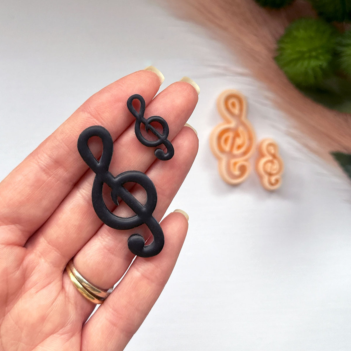 A hand holds two black treble clef charms, with two peach-colored treble clef clay cutters blurred in the background on a white surface.