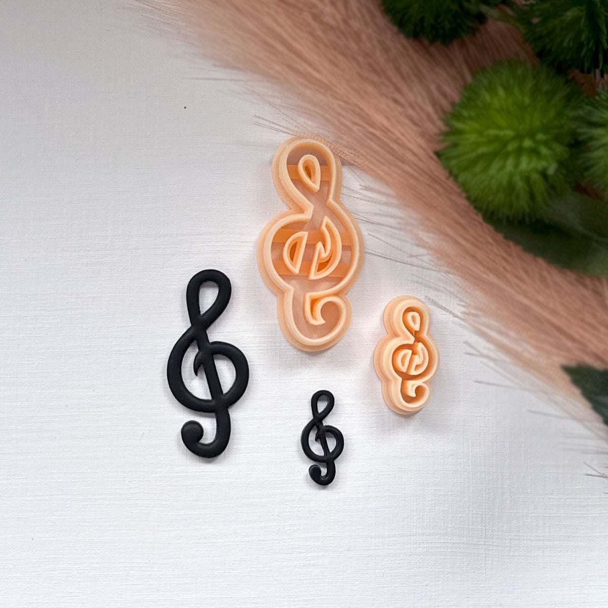 Two treble clef-shaped clay pieces in black and 2 peach-colored pla clay cutters, arranged on a white textured surface with a soft beige grass and green foliage in the background.
