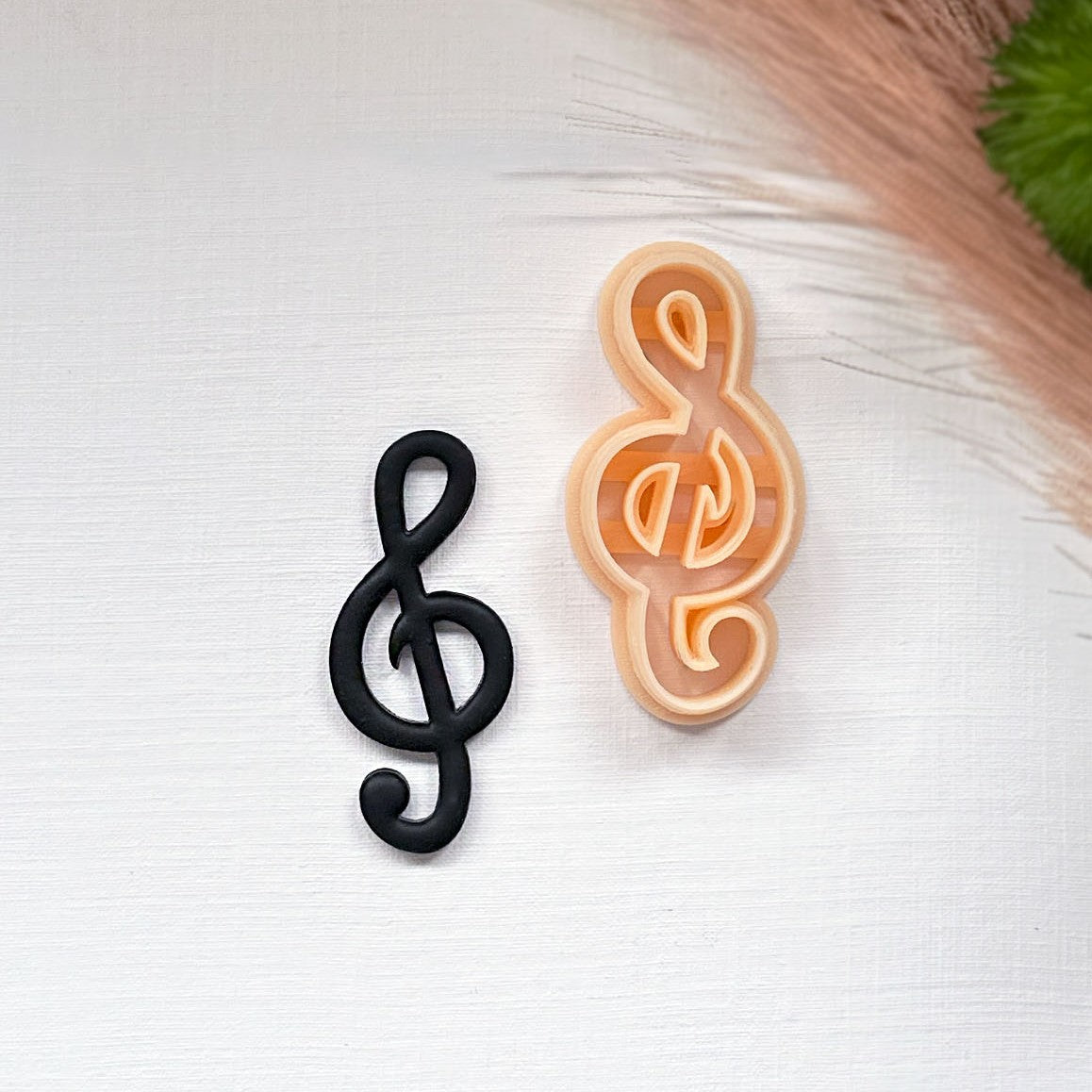 Two treble clef-shaped items on a white textured surface: one matte black clay cutout and one peach-colored plastic clay cutter, with dried grass and a green leaf in the top right corner.