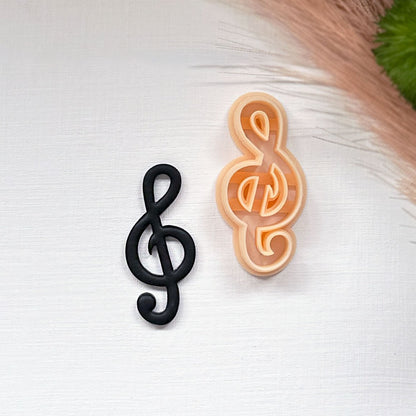 Two treble clef-shaped items on a white textured surface: one matte black clay cutout and one peach-colored plastic clay cutter, with dried grass and a green leaf in the top right corner.
