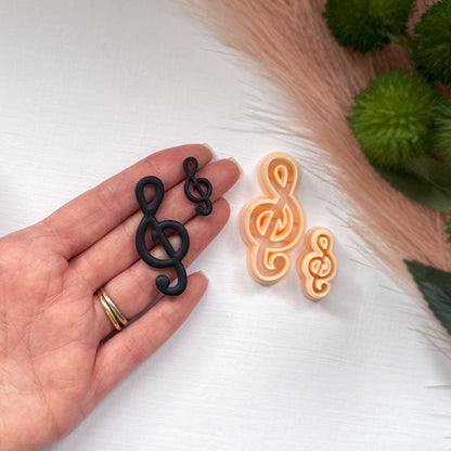 A hand holds two black treble clef-shaped items next to two peach-colored treble clef-shaped clay cutters on a white surface with decorative grass and greenery.