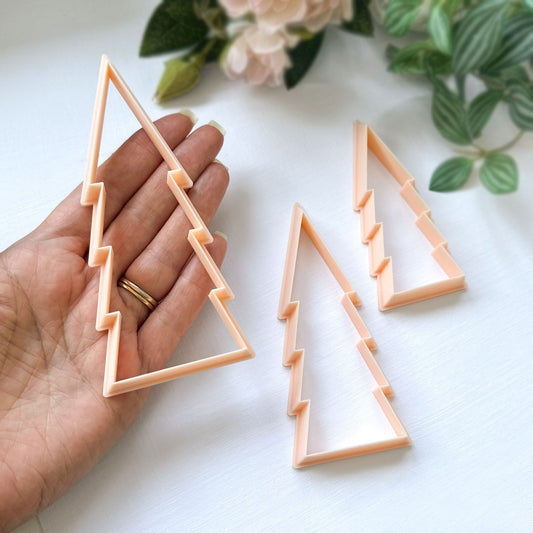 Three peach-colored plastic cookie clay cutters shaped like stylised Christmas trees, with one being held in a hand to show scale on a white surface with soft floral background. Great for ornament making.