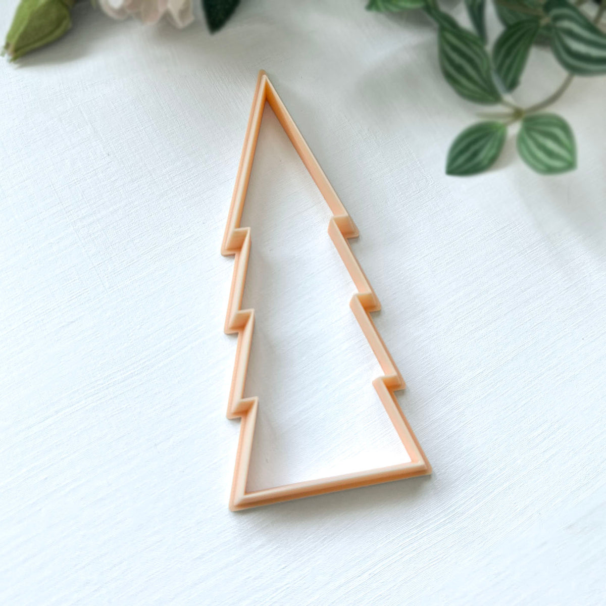 A peach-colored plastic cookie clay cutters shaped like a stylised Christmas tree, on a white surface with soft floral background. Great for ornament making.
