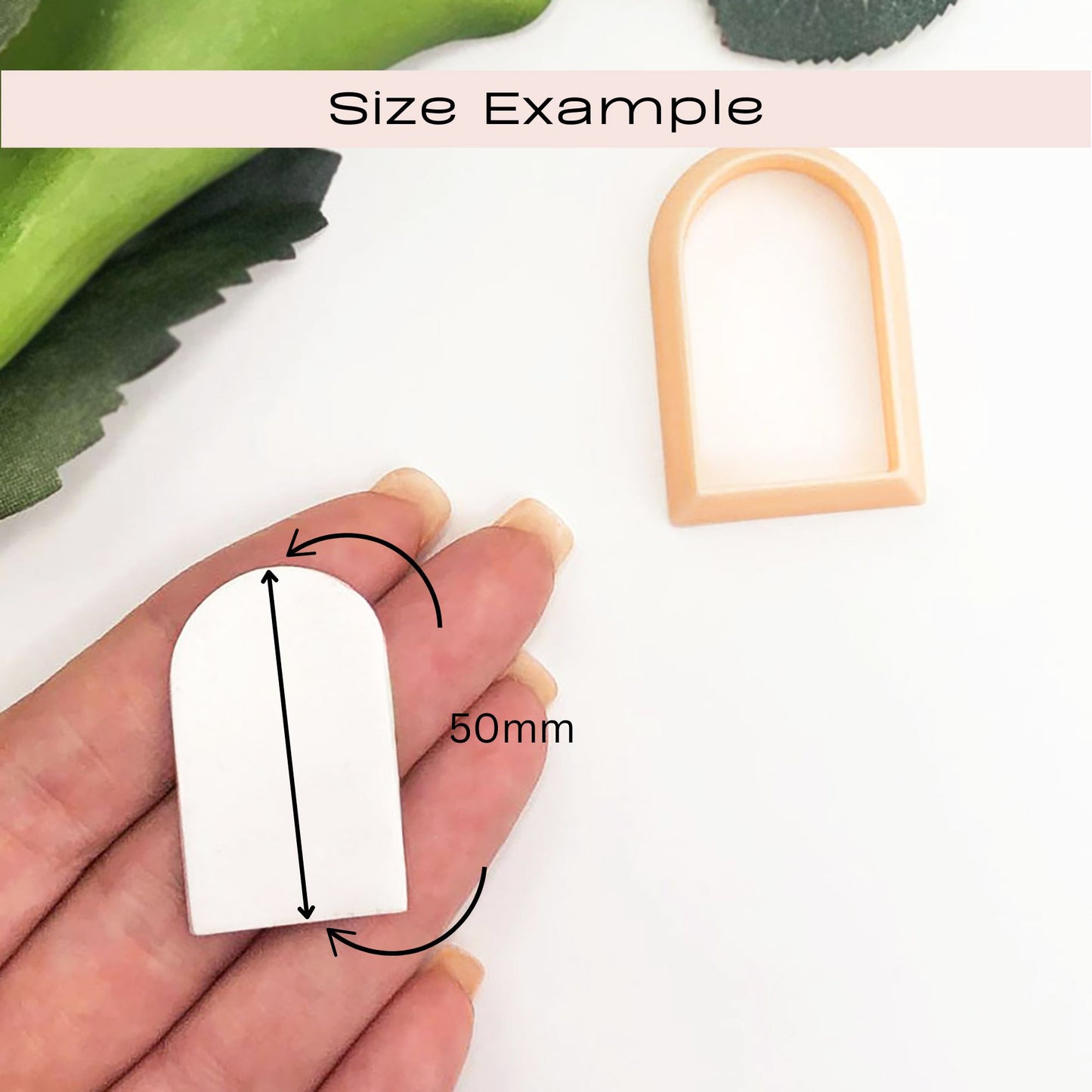 Hand holding a white polymer clay arch with measurement details, set against a white background with green leaves.