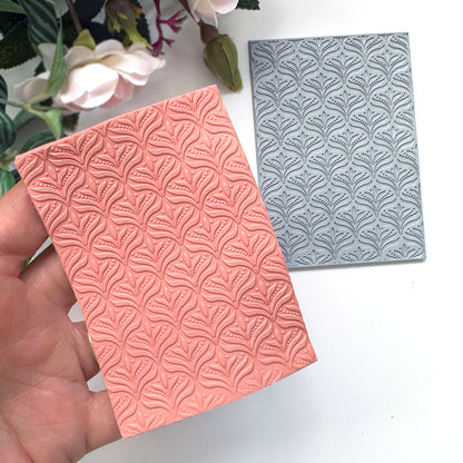A hand holds a rectangular coral-pink embossed polymer clay slab with a repeating leaf-like floral pattern, while a matching gray embossed rubber stamp is placed beside it on a white surface with flowers in the background.