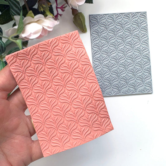 A hand holds a rectangular coral-pink embossed polymer clay slab with a repeating leaf-like floral pattern, while a matching gray embossed rubber stamp is placed beside it on a white surface with flowers in the background.
