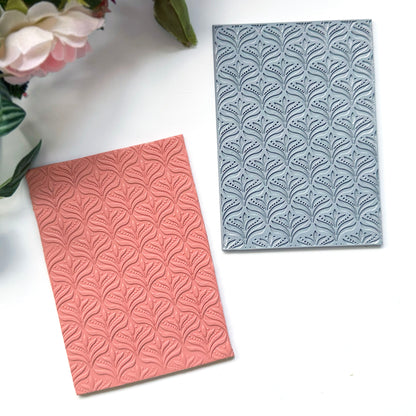 A grey rectangular embossed rubber stamping mat with a repeating floral leaf pattern, shown on a white background. Next to it is a coral polymer clay slab with the pattern stamped in it, with decorative flowers in the top-left corner.