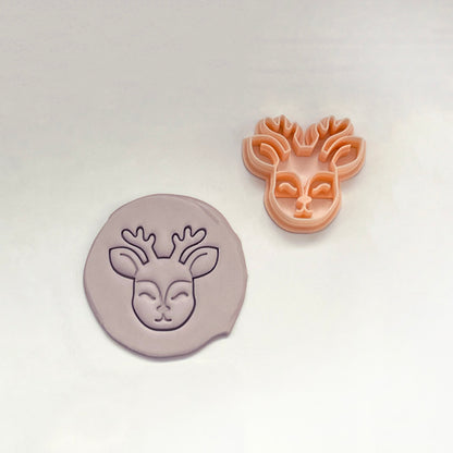 A deer head cookie cutter in peach-colored plastic and a matching circular clay stamp in a dusty lavender-purple, displayed on a clean white background.