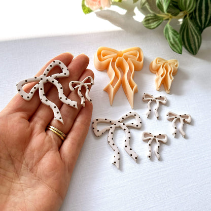Hand holding bow-shaped polymer clay cut-outs on a light surface with the clay cutters in the front and a plant in the background