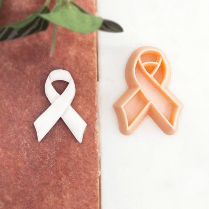 A white awareness ribbon clay cut out and a peach-colored awareness ribbon cookie cutter are displayed on a split brown and white background with a green leaf in the corner.