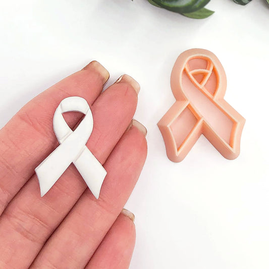 A small white awareness ribbon polymer clay charm is held on fingers next to a peach-colored awareness ribbon clay cookie cutter on a white background.