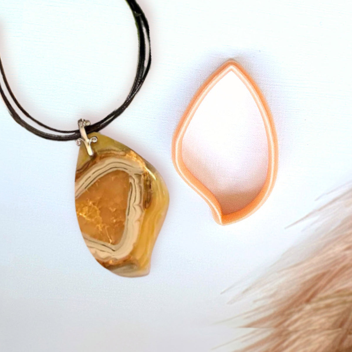 A teardrop-shaped pendant necklace made from a faux polished agate slice of polymer clay hanging on a black cord, shown next to its peach-colored PLA 3D printed clay cutter on a white background.